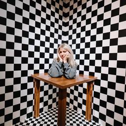 Persoon zit aan een tafel in een kamer met zwart-witte schaakbordpatronen die een optische illusie creëren.