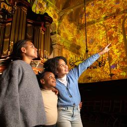 Drie bezoekers kijken omhoog naar kleurrijke projecties van kunstwerken op de muren en het plafond van een historische kerkruimte.